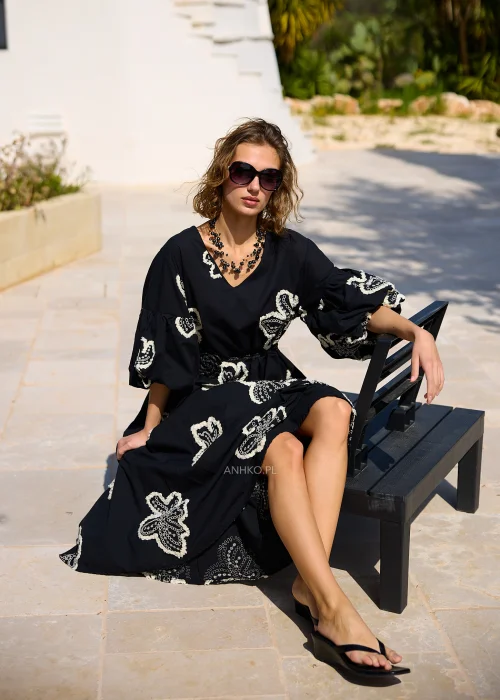 Czarna sukienka maxi z efektownymi białymi haftami w kształcie kwiatów, bufiastymi rękawami i ozdobnym paskiem w talii. Zwiewny model w stylu boho, idealny na eleganckie i codzienne stylizacje z kobiecym charakterem.