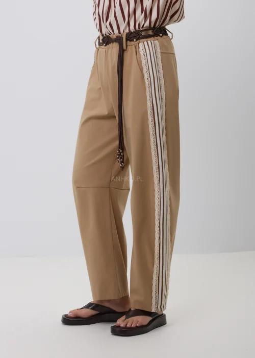 Beżowe damskie spodnie o luźnym fasonie z szerokimi nogawkami i ozdobnym koronkowym lampasem po bokach. Stylowy model casual z elastycznym pasem, szlufkami i plecionym paskiem w odcieniach brązu.