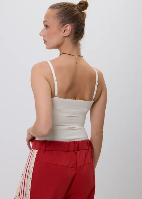 Kremowy top damski na cienkich regulowanych ramiączkach z wyjmowanymi wkładkami, o dopasowanym kroju i minimalistycznym designie, prezentowany w stylizacji z czerwonymi spodniami.