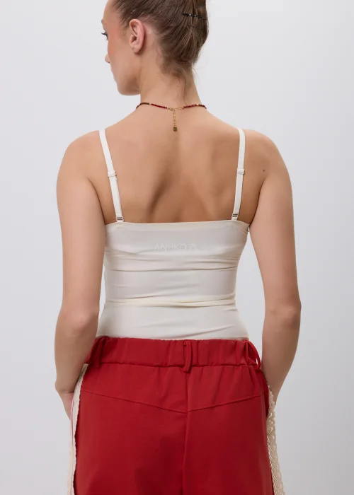 Kremowy top damski na cienkich regulowanych ramiączkach z wyjmowanymi wkładkami, o dopasowanym kroju i minimalistycznym designie, prezentowany w stylizacji z czerwonymi spodniami.