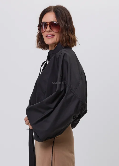 Czarna damska kurtka bomber oversize ze stójką, szerokimi rękawami i regulowanymi troczkami przy mankietach. Nowoczeska stylizacja casual z krótką kurtką i beżową spódnicą maxi.