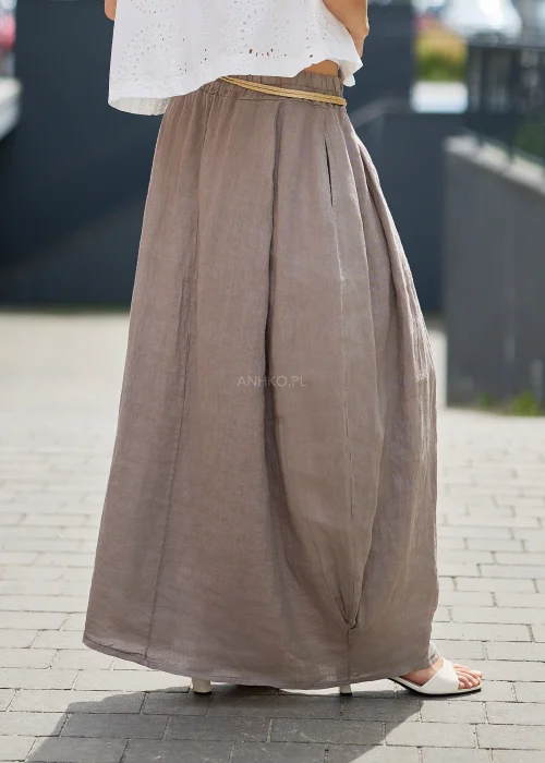Brązowa lniana spódnica maxi o luźnym fasonie z elastyczną gumką w pasie i ozdobnym plecionym paskiem, prezentowana w stylizacji z białym topem i ażurową kamizelką.