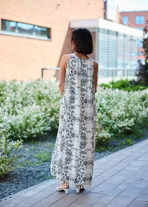 Beżowa sukienka maxi damska z czarnym roślinnym wzorem, dekoltem halter i ozdobnym kwiatem przy ramieniu. Zwiewny fason w stylu boho wykończony chwostami, zestawiony z plecioną brązową torebką i letnimi dodatkami.