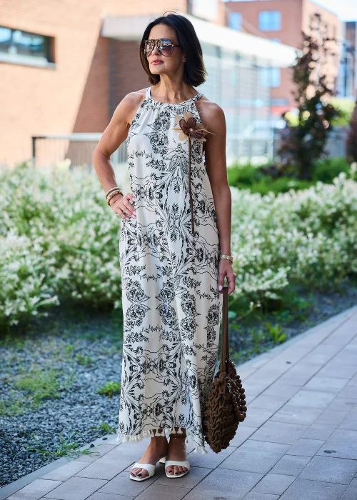 Beżowa sukienka maxi damska z czarnym roślinnym wzorem, dekoltem halter i ozdobnym kwiatem przy ramieniu. Zwiewny fason w stylu boho wykończony chwostami, zestawiony z plecioną brązową torebką i letnimi dodatkami.