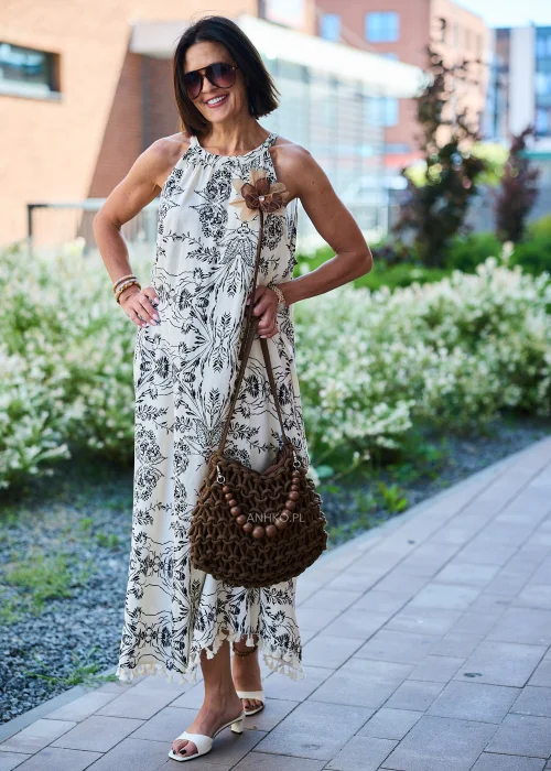 Beżowa sukienka maxi damska z czarnym roślinnym wzorem, dekoltem halter i ozdobnym kwiatem przy ramieniu. Zwiewny fason w stylu boho wykończony chwostami, zestawiony z plecioną brązową torebką i letnimi dodatkami.