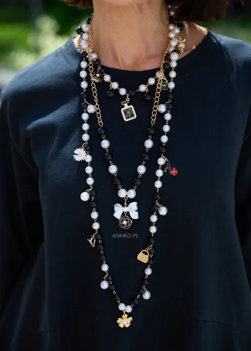 Długi naszyjnik z czarnych i białych pereł oraz złotego łańcuszka, ozdobiony eleganckimi zawieszkami w kształcie kwiatów, kokardki, flakonika perfum, szpilki i listka.