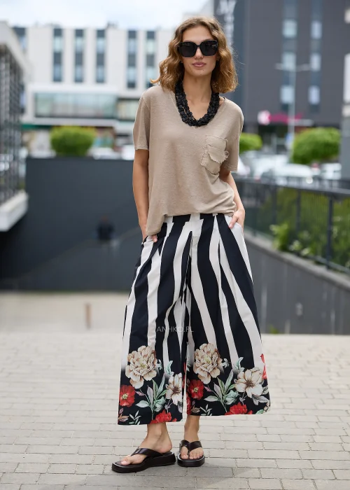 Beżowy t-shirt z dekoltem V i postrzępioną kieszonką na piersi. Luźny model zestawiono z czarno-białymi spodniami w szerokie pasy oraz czarnym, wielorzędowym naszyjnikiem, tworząc nowoczesną stylizację casual.